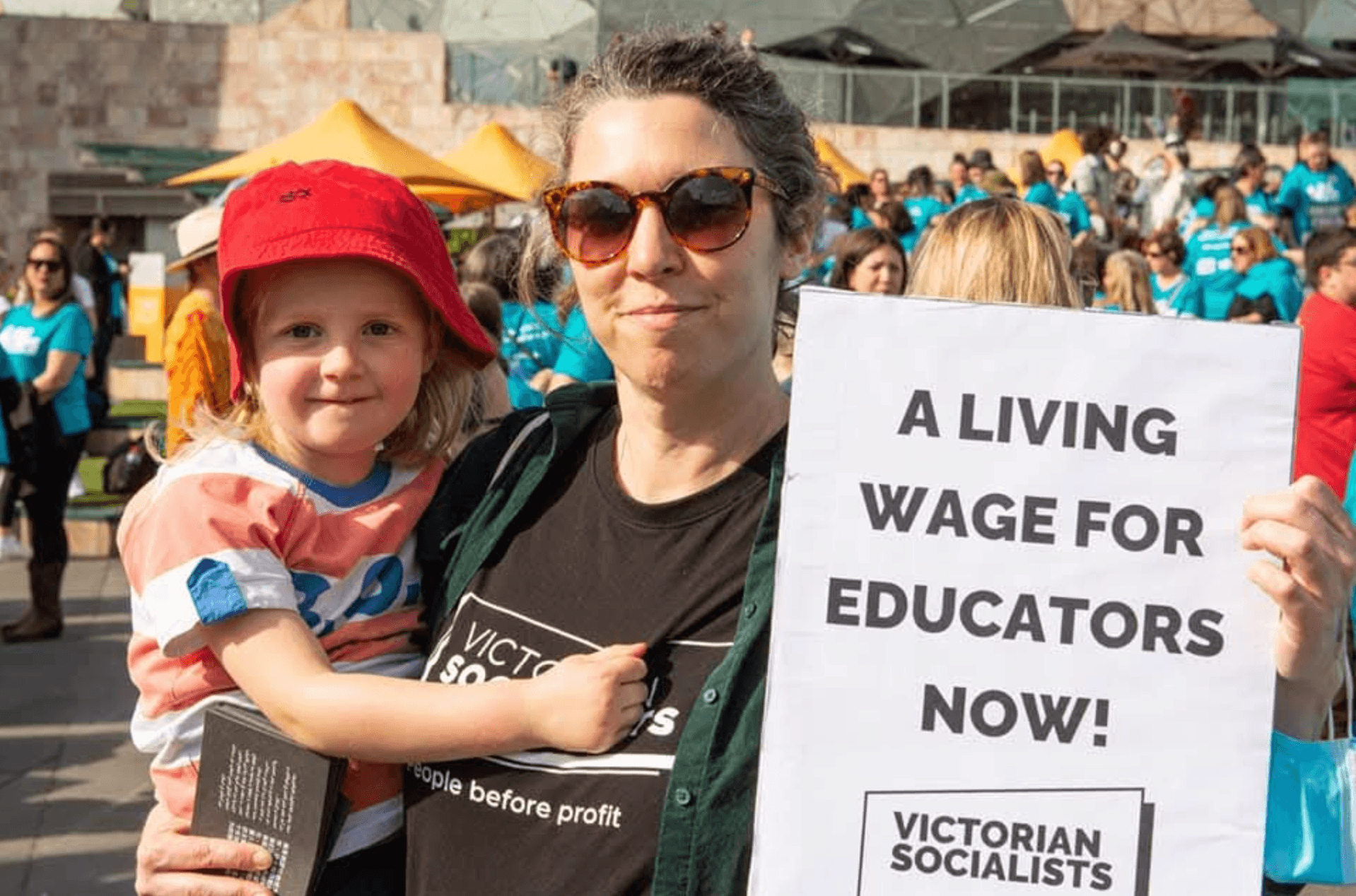 A woman holds a toddler in one arm and a sign that reads 'A living wage for educators now!' with the VS logo in the other